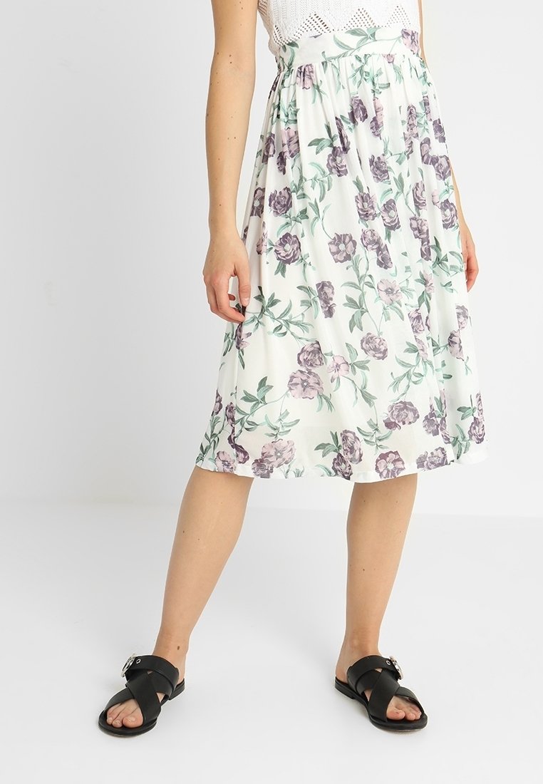 Femme portant une jupe blanche fleurie longueur genou avec des fleurs vertes et violettes et des sandales noires à brides croisées debout sur un sol blanc.