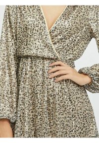 Klänning med leopardmönster som har V-hals, långa puffärmar och vikt midja, prydd med glittrande gyllene paljetter på en ljusbeige bakgrund.