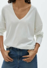 Maglione bianco a collo a V con maniche lunghe, texture morbida, vestibilità rilassata, abbinato a jeans di denim blu. Nessun motivo o accento visibile.
