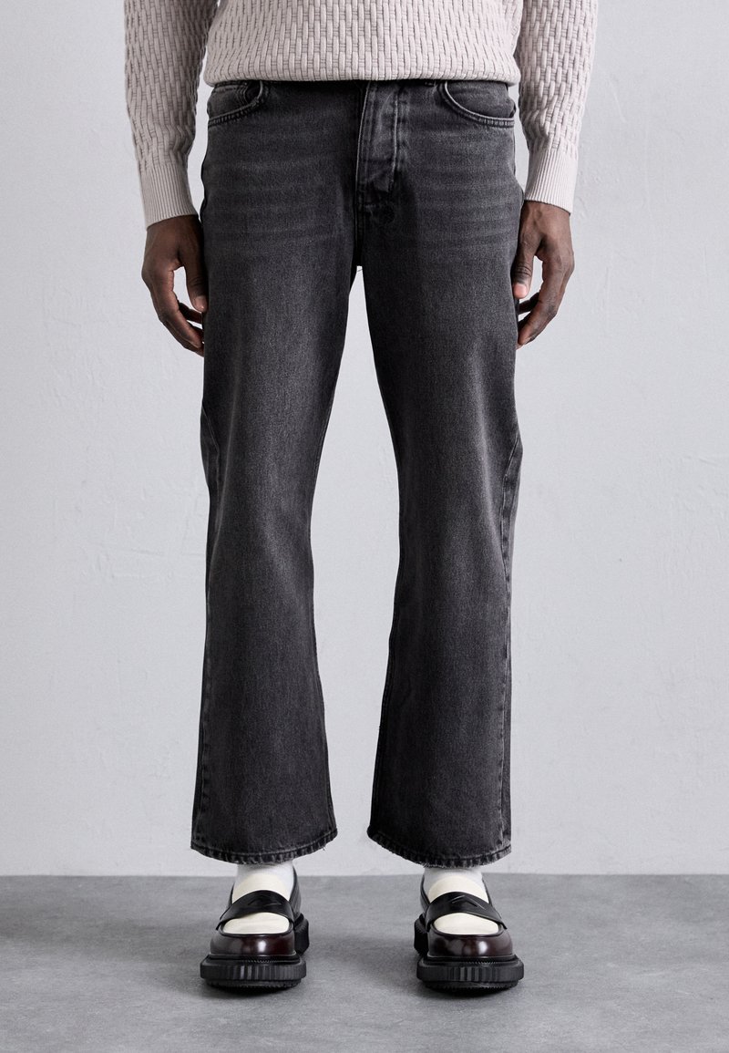 Jeans en denim noir avec une coupe droite et cropped, légèrement délavés, et un design à cinq poches, portés avec des chaussures noires et blanches.