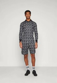 Vīrietis stāv, saskatījies uz priekšu, tērpies saskaņotā melnā un pelēkā ģeometrisko rakstu garajās piedurknēs kreklā un šortos, melnās kurpēs un baltos svītrainos zeķēs.