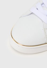 Hvit sneakers med et teksturert lærøvre, med en gullaccent langs sålen og hvite snørebånd. Nærbilde av tåområdet.