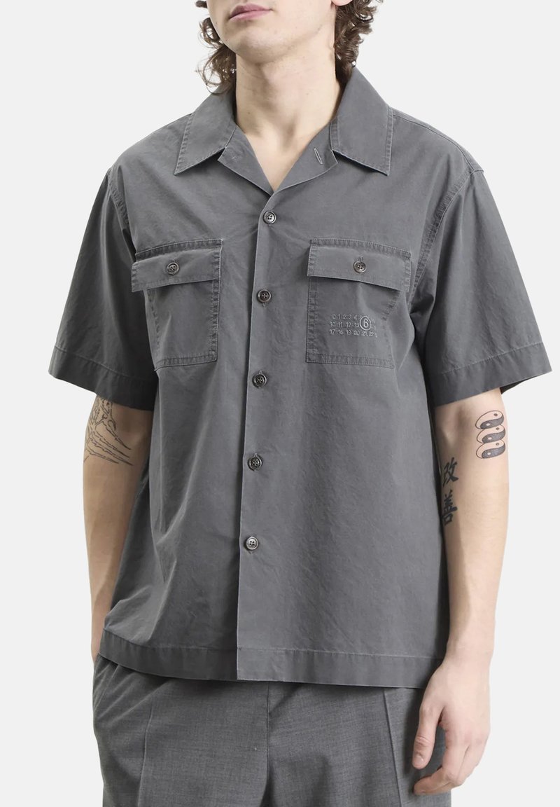 Uomo che indossa una camicia grigia a maniche corte e corte, con due tasche sul petto, in piedi con le mani nelle tasche dei pantaloni grigi, tatuaggi visibili sulle braccia.