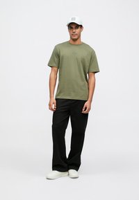 Maglietta verde oliva a maniche corte, con un logo sottile al centro. Abbinata a pantaloni neri a gamba larga e scarpe da ginnastica bianche.