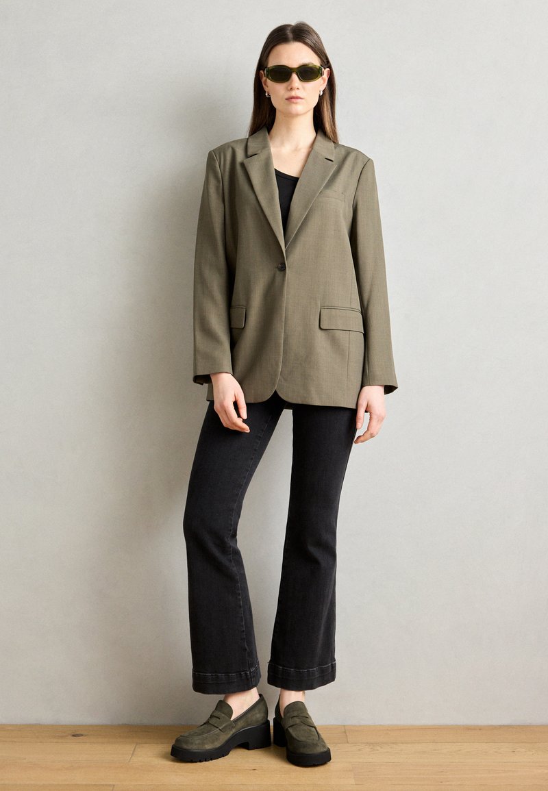 Blazer verde oliva, top nero, jeans neri svasati e scarpe verde con suola spessa; occhiali da sole verdi oversize completano il look.