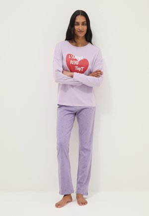 Woman stands barefoot with arms crossed wearing lavender pajamas; top has a red heart and French text "L'amour rend tout."