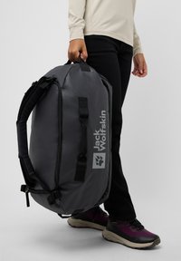 Bolsa de viaje gris con una textura suave, con correas y herrajes negros. Tiene un logo prominente, cierre de cremallera y un diseño espacioso.