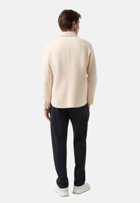 Jasno beżowa koszula typu kurtka z wełny z kołnierzem, z prostym szwem na plecach, w połączeniu z granatowymi spodniami i białymi trampkami.