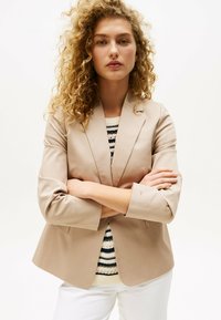 Blazer beige su misura con rever appuntito, chiusura a un bottone e maniche a tre quarti, indossato sopra un maglione a righe.