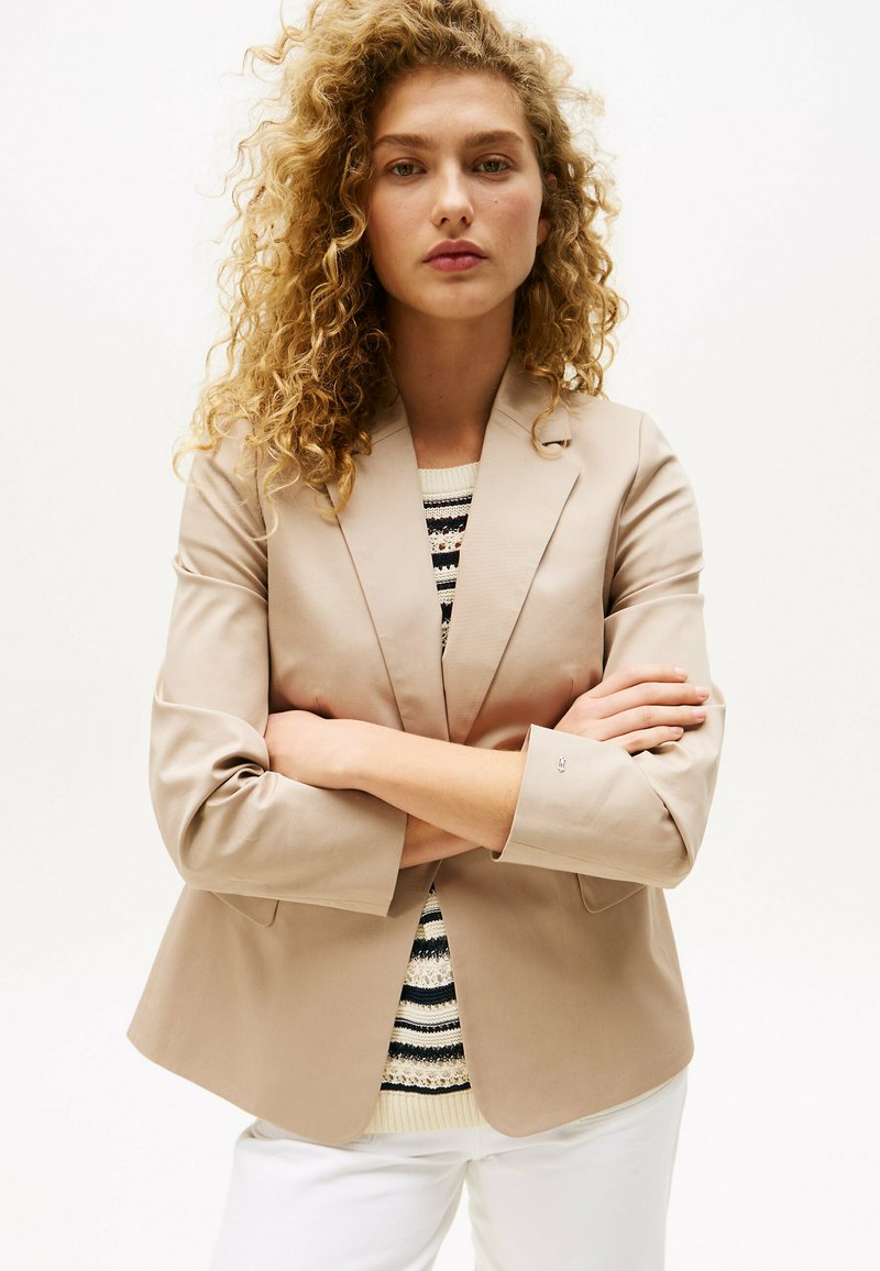 Blazer beige su misura con rever appuntito, chiusura a un bottone e maniche a tre quarti, indossato sopra un maglione a righe.