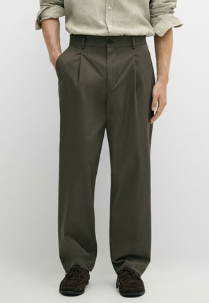 Mann trägt weite dunkelolivfarbene Hose mit Falten und schwarze Schnürschuhe, Hände in den Taschen, kombiniert mit einem leichten Hemd mit hochgekrempelten Ärmeln.