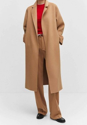 Person iført en lang kamelfarvet frakke, matchende kamelfarvede bukser, en rød top og sorte sko, stående mod en ensartet lys baggrund.