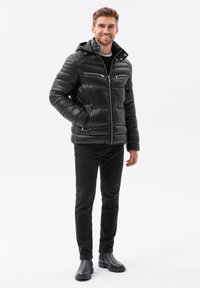 Chaqueta negra acolchada con capucha, bolsillos con cremallera y un acabado brillante, combinada con jeans negros y botas negras hasta el tobillo.