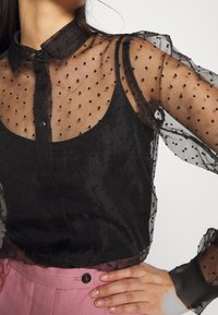 Blouse noire transparente à motif à pois, avec fermeture boutonnée sur le devant et col. Dotée de manches longues bouffantes et de textures superposées.
