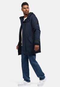 Parka longue bleu marine avec fermeture éclair et bouton, capuche ajustable, poches latérales et poignets côtelés. Col haut avec une doublure intérieure douce.