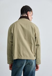 Olijfgroene katoenen jas met een bruine corduroy kraag, relaxte pasvorm, drukknopen en zichtbare stiksels. Ook worden blauwe jeans gedragen.