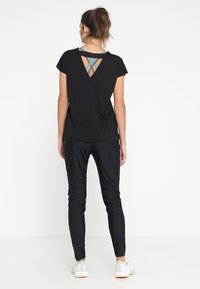 Czarna sportowa koszulka z wycięciem na plecach, w połączeniu z eleganckimi czarnymi spodniami i białymi trampkami. Materiał wydaje się lekki i przewiewny.