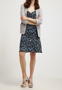 Schwarzes Blumenkleid mit blauen und weißen Blumen, kombiniert mit einem hellgrauen, strukturierten Blazer. Beiger Plateau-Absatz rundet das Outfit ab.
