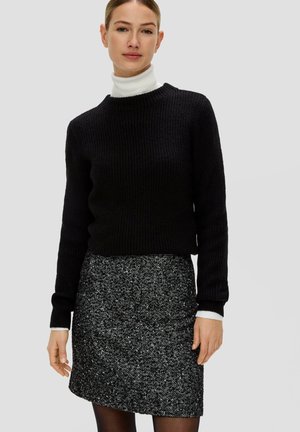 Schwarzer gerippter Strickpullover mit hohem Kragen, kombiniert mit einem strukturierten schwarzen und weißen gepunkteten Minirock, der kontrastierende Bündchen zeigt.