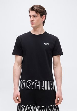 Jonge man die een zwarte Moschino T-shirt en korte broek draagt met groot wit logo, kijkend naar links tegen een eenvoudige achtergrond.