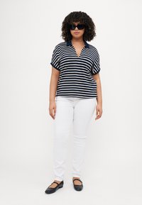 Polo a righe blu navy e crema con scollatura a V e maniche corte, abbinato a jeans bianchi e scarpe flat nere. Gli accessori includono occhiali da sole.