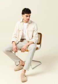 Beige raidoitu paita valkoisen T-paidan päällä, vaaleanharmaat pellavahousut ja ruskeat mokkanahkaliukupohjakengät; istuu metallikehyisessä puutuolissa.