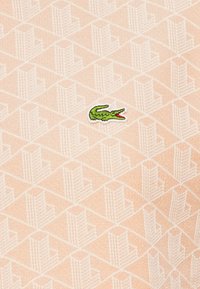 Kartotas šviesiai rožinis geometrinis raštas su mažu žaliu siuvinėtu krokodilo logotipu, esančiu audinio centre.