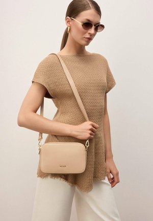 Vrouw draagt beige gebreide top en witte broek, met een beige crossbodytas, een zonnebril en gouden oorringen.