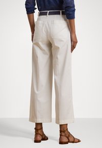 Pantalons larges blancs avec poches arrière, en coton, assortis d'une ceinture beige et de sandales marron, montrant une coupe décontractée et une texture lisse.