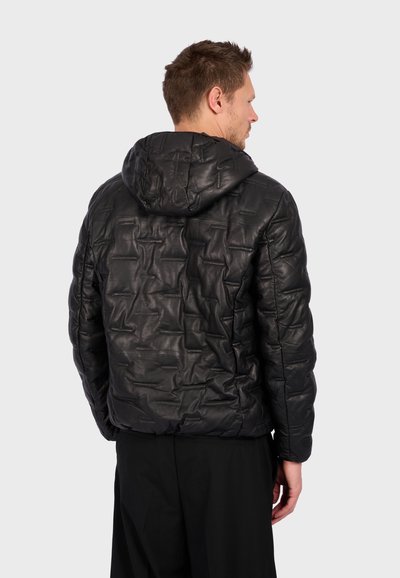 Homme de dos portant une veste à capuche noire matelassée avec un motif texturé et un pantalon noir devant un fond clair.