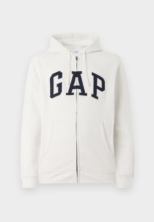 Sweat à capuche zippé couleur crème en tissu doux, arborant un grand logo "GAP" en marine sur le devant, avec deux poches avant et une capuche.