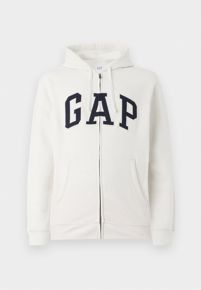 Kremowy hoodie z zamkiem wykonany z miękkiego materiału, z dużym granatowym logo "GAP" z przodu, dwoma kieszeniami z przodu oraz kapturem.