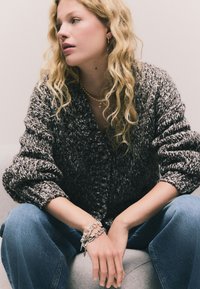 Adīts kardigans pelēkā un baltā krāsā ar rupju tekstūru un lielām pogām. Kombinēts ar zilām džinsa biksēm un sudraba ķēdes aproci.