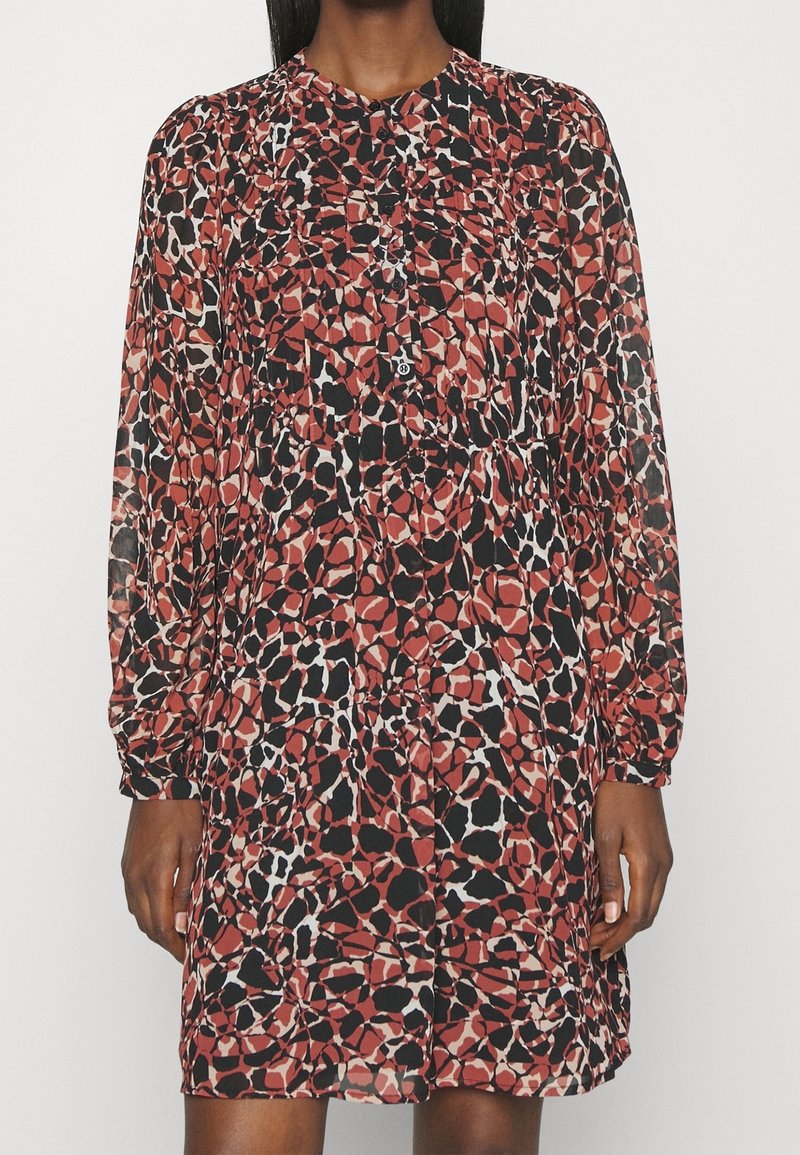 Femme portant une robe à manches longues, longueur genou, avec un motif léopard abstrait rouge, noir et blanc et des détails de boutons sur le devant.