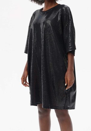 Frau trägt ein locker sitzendes schwarzes Paillettenkleid mit ellbogenlangen Ärmeln, steht mit entspannten Armen vor einem weißen Hintergrund.