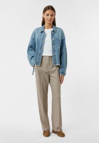 Giacca di denim azzurro chiaro con chiusura a bottoni, t-shirt bianca sotto, pantaloni a quadri beige e scarpe in suede marroni.