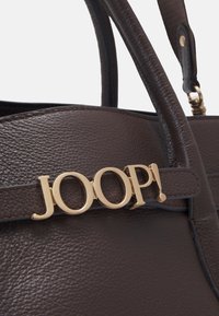 Brązowa skórzana torebka o fakturowanym wykończeniu, z ozdobą w postaci złotego logo "JOOP!" na pasującym pasku. Uchwyt zaprojektowany z myślą o wygodnym noszeniu.
