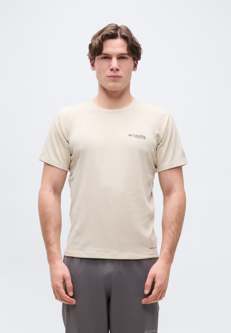 Νέος άνδρας που φοράει μπεζ κοντομάνικο πουκάμισο Columbia Titanium και γκρι παντελόνι, στέκεται μπροστά σε απλό φωτεινό φόντο.