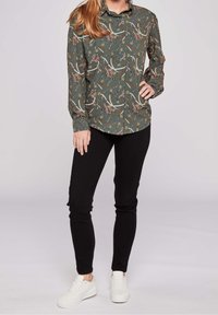 Ciemnozielona koszula z długim rękawem w kwiatowy i zwierzęcy wzór, zestawiona z czarnymi spodniami slim-fit i białymi tenisówkami.