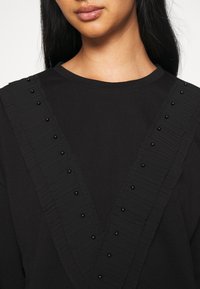 Haut noir avec un décolleté en V orné de volants et de détails en perles noires le long des bords. Tissu lisse et texturé.