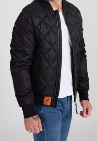 Blouson aviateur noir matelassé avec poignets côtelés, fermeture éclair à l'avant et détail d'étiquette orange à l'ourlet. Porté par-dessus une chemise blanche et un jean en denim.