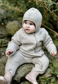 Sive boje dječji kombinezon od flisa s prednjim patentnim zatvaračem, kapuljačom i rebrastim detaljima; uparen s ružičastim gumama. Sjedi na stijeni.