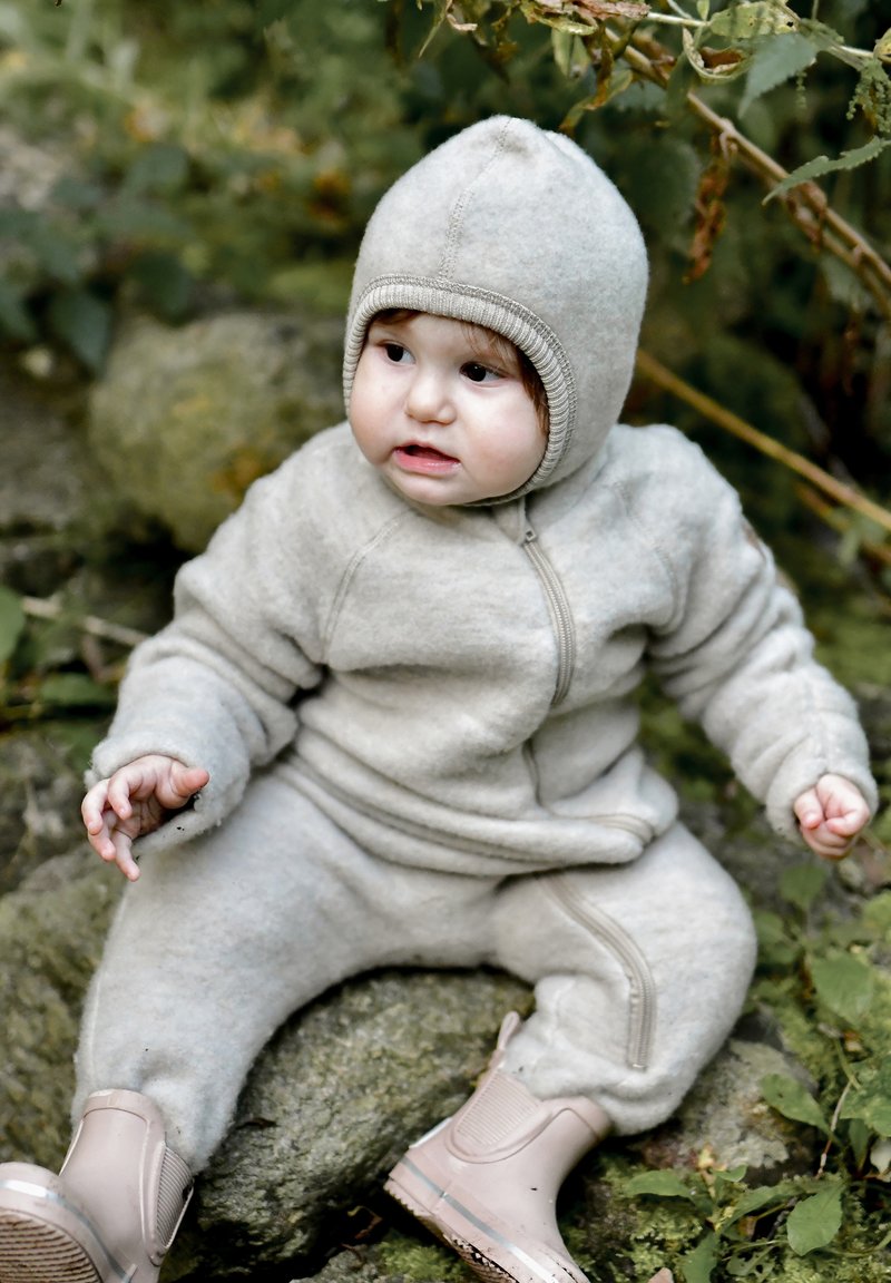 Sive boje dječji kombinezon od flisa s prednjim patentnim zatvaračem, kapuljačom i rebrastim detaljima; uparen s ružičastim gumama. Sjedi na stijeni.