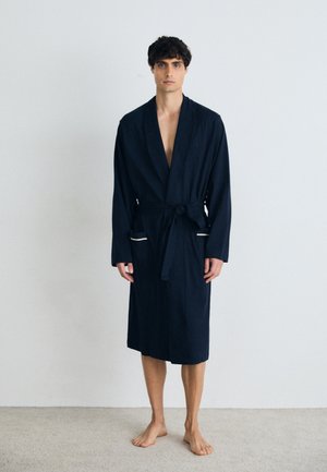 Accappatoio in spugna blu navy con scollo a V profondo, maniche lunghe, vita con cintura e tasche frontali con accenti crema. Texture liscia, design lungo fino al ginocchio.