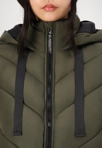 Kobieta ubrana w oliwkowo-zieloną pikowaną kurtkę z czarnym zamkiem błyskawicznym i sznurkami od kaptura, widoczne usta, tło jednolicie białe.