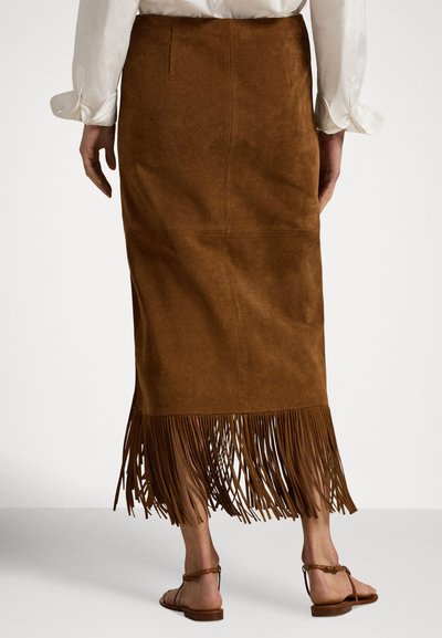 Ženska, oblečena v rjavo semiš krilo do sredine meč s franžami na robu in belo dolgo srajco z dolgimi rokavi, stoji bosa na belem tlaku.