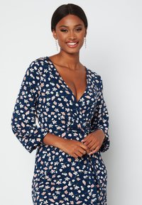 Granatowa sukienka w kwiaty z głębokim dekoltem w serek, długimi rękawami i związaną talią, zdobiona małymi różowymi i białymi wzorami kwiatowymi.