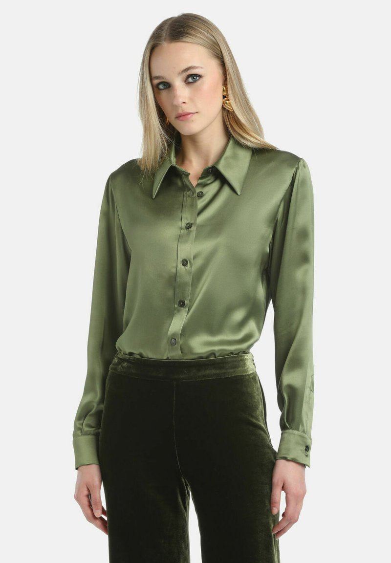 Satinasta zelena bluzica s klasičnom ovratnikom, dugim rukavima i zatvaranjem na dugmad. Nosila se s baršunastim zelenim hlačama. Glatka tekstura i elegantan završetak.