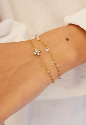 Zwei filigrane goldene Gliederarmbänder mit kleinen Kristallanhängern schmücken ein Handgelenk mit sichtbarem Muttermal vor einem hellen Stoffhintergrund.