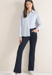 Chemise à rayures bleu clair et blanc à boutons, associée à un jean évasé bleu foncé, avec une poche latérale et des coutures contrastantes. Baskets blanches.